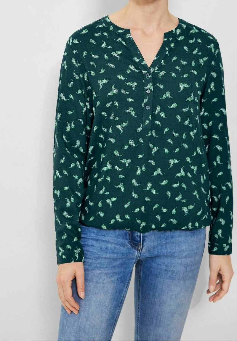 Femme portant une chemise à manches longues vert foncé avec des motifs paisley vert clair et un jean bleu, debout devant un fond uni.