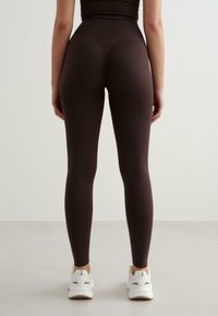 Asmuo dėvi juodas, aukštai juosmeniu priglundančias leggings ir baltus sportbačius, stovintis atsukęs nugara prieš paprastą šviesią foną.