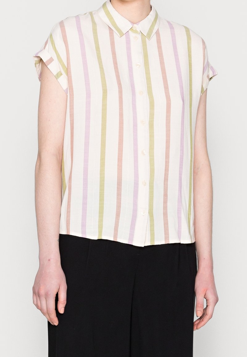 Femme portant une chemise à manches courtes boutonnée avec des rayures pastel verticales en vert, violet et beige, assortie à un pantalon noir.