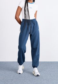 Modré tepláky s elastickým pasem a manžetami, s detailem loga Nike; doplněné o bílé tričko a bílé tenisky.