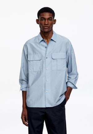 Muž nosiaci svetlomodrú košeľu na zapínanie s dvoma náprsnými vreckami a vyhrnutými rukávmi, v kombinácii s tmavými nohavicami, stojaci pred bielym pozadím.
