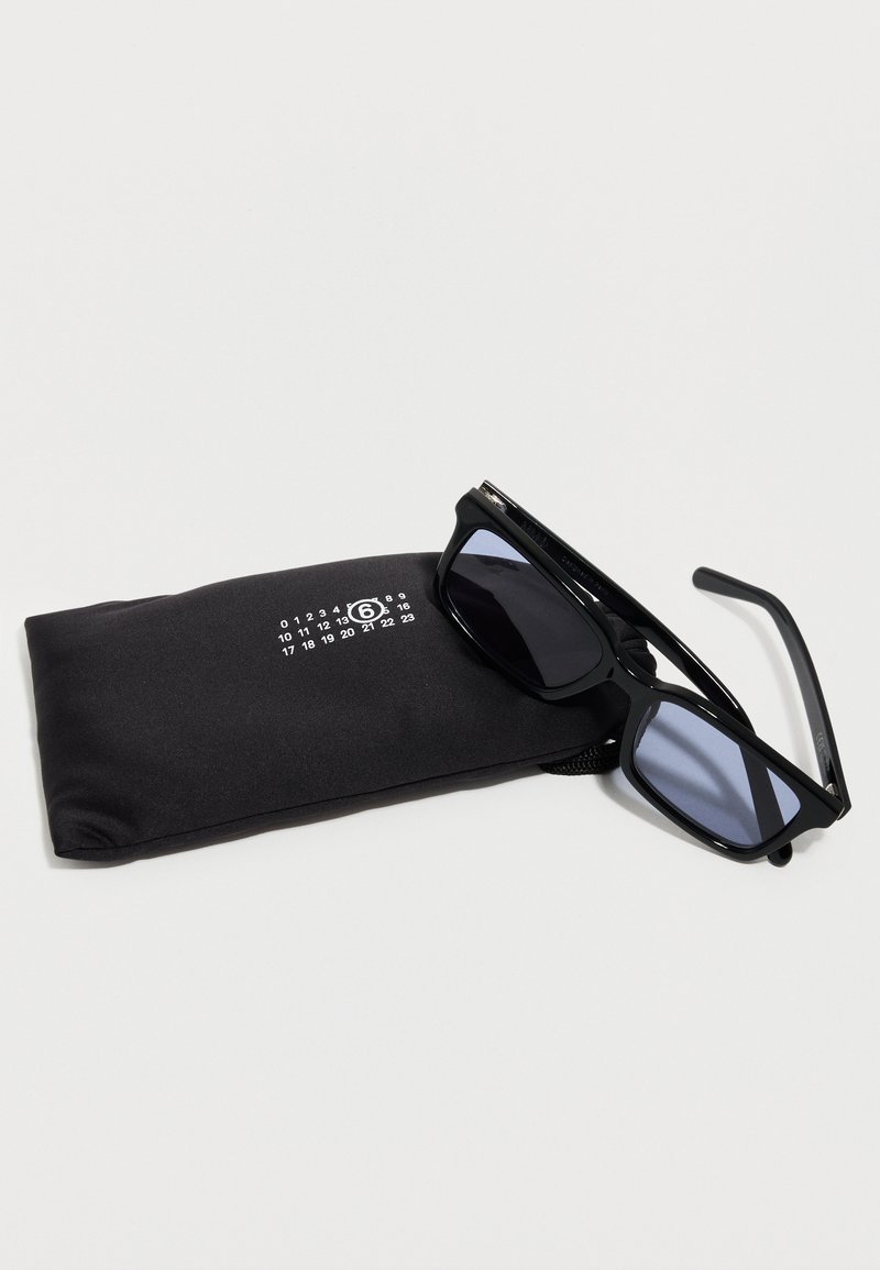 Lunettes de soleil rectangulaires noires posées sur une pochette en tissu noire avec un logo numérique blanc sur un fond blanc.