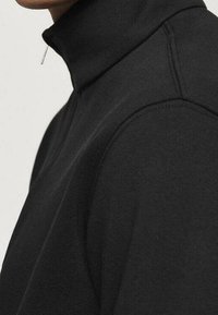 Felpa nera con collo alto, realizzata in tessuto morbido. Presenta una zip al collo, una consistenza liscia e maniche raglan. Dettagli di cuciture minimi.