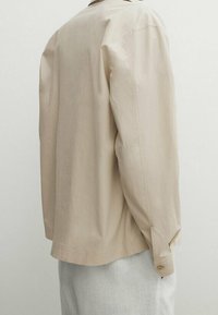 Camicia beige chiaro con una vestibilità rilassata, dotata di polsini abbottonati e colletto semplice. Tessuto liscio, design minimalista, taglio dritto.