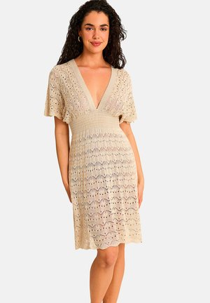 Femme aux cheveux bouclés portant une robe tricotée beige avec un profond décolleté en V, des manches courtes et une taille cintrée, debout devant un fond uni.