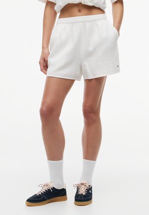 Kobieta ubrana w białe teksturowane szorty, białe skarpetki do kostki oraz granatowe sneakersy z gumowymi podeszwami, stojąca z jedną ręką w kieszeni.