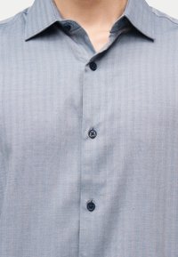 Κοντινό πλάνο ενός ατόμου που φοράει πουκάμισο με σχέδιο ψαροκόκκαλο σε ανοιχτό γαλάζιο χρώμα, με σκούρα μπλε κουμπιά, γιακά και ανοιχτό το πάνω κουμπί.