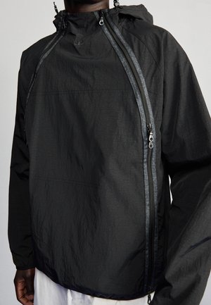 Veste légère noire avec zip étanche en diagonale et capuche portée par une personne à la peau foncée et pantalon blanc.