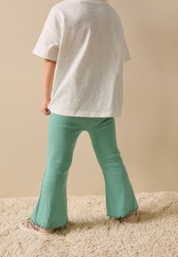 Könnyű, fehér pamutpóló, mentazöld bordázott, bővülő szárú nadrággal, nyers szegéllyel, bézs tornacipővel viselve.