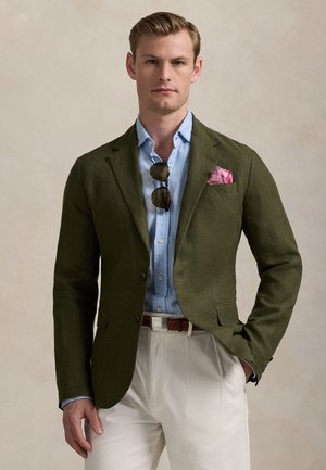 Man står självsäkert i en olivgrön blazer över en blå skjorta, med en rosa fickduk och solglasögon, mot en neutral bakgrund.