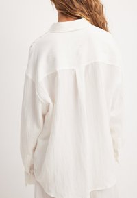 Valkoinen pitkähiha shirt, jossa on sileä ja hieman teksturoitu kangas. Ominaisuuksiin kuuluvat klassinen kaulus ja napilliset hihansuut, rento istuvuus ja kaareva helma.