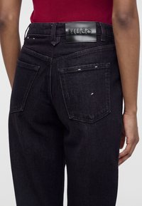 Personne portant un jean en denim noir avec des poches arrière et une étiquette en cuir noir embossée avec "HUGO" sur la ceinture.