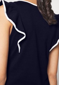 Tmavě modrý top s krátkými rukávy a volánkovými rukávy s bílým lemováním. Měkký bavlněný materiál s hladkou texturou a klasickým střihem.