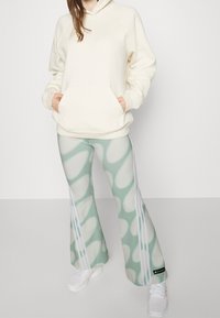 Cremehuvtröja med framficka, matchad med gröna mönstrade utsvängda byxor med vita ränder och en svart logotypdetalj. Vita sneakers.