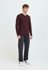 Burgunderfarget V-hals-genser i et mykt strikket materiale, kombinert med mørkegrå jeans og hvite joggesko med svarte striper, båret av en stående mannlig modell.
