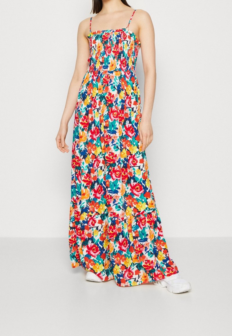 Robe maxi à fines bretelles, présentant un motif floral coloré sur un fond clair, corsage smocké et jupe à volants.