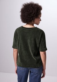 Top corto de color verde oscuro con textura, de ajuste relajado y cuello redondo, combinado con unos jeans de mezclilla azules. La vista trasera muestra las mangas enrolladas.