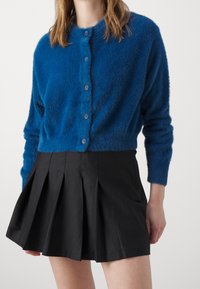 Niebieski, puszysty kardigan z okrągłym dekoltem i pięcioma pasującymi guzikami, noszony na czarnej plisowanej spódnicy. Miękka tekstura, długie rękawy.