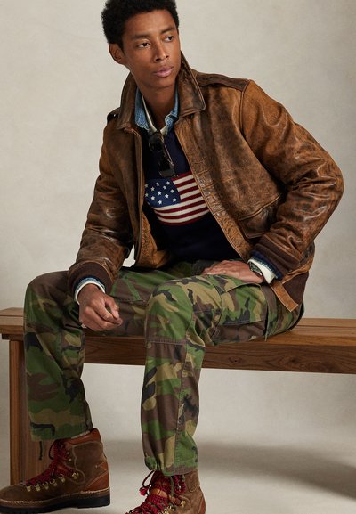 Bruine leren jas over een blauw sweater met een ontwerp van de Amerikaanse vlag, camouflage cargobroek en bruine laarzen met rode veter, zittend op een houten bank.