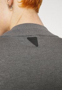 Suéter gris de canalé con escote redondo, que presenta una etiqueta triangular de tela negra en la parte posterior cerca del cuello. Textura suave.