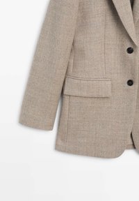 Lichtbeige blazer van textuurstof, met een enkele borstzak, ingeknipte revers en drie zwarte knopen aan de voorkant.