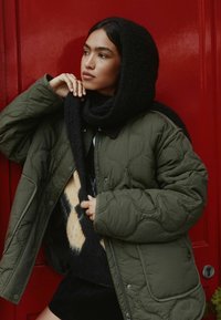 Gestepptes olivgrünes Jackett mit einem schwarzen Strickschal. Darunter trägt die Person einen gemusterten Pullover mit beigen und marineblauen Akzenten. Der Hintergrund ist eine rote Wand.