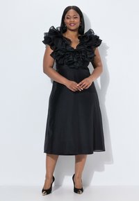Robe noire avec un corsage ajusté et de grandes manches à volants, en tissu texturé ; longueur genou avec une jupe évasée et des chaussures à talons hauts.