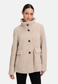 Beżowy, teksturowany płaszcz z podniesionym kołnierzem, z pięcioma czarnymi guzikami i dwoma przednimi kieszeniami. Elegancki, przytulny krój z miękkiego materiału.