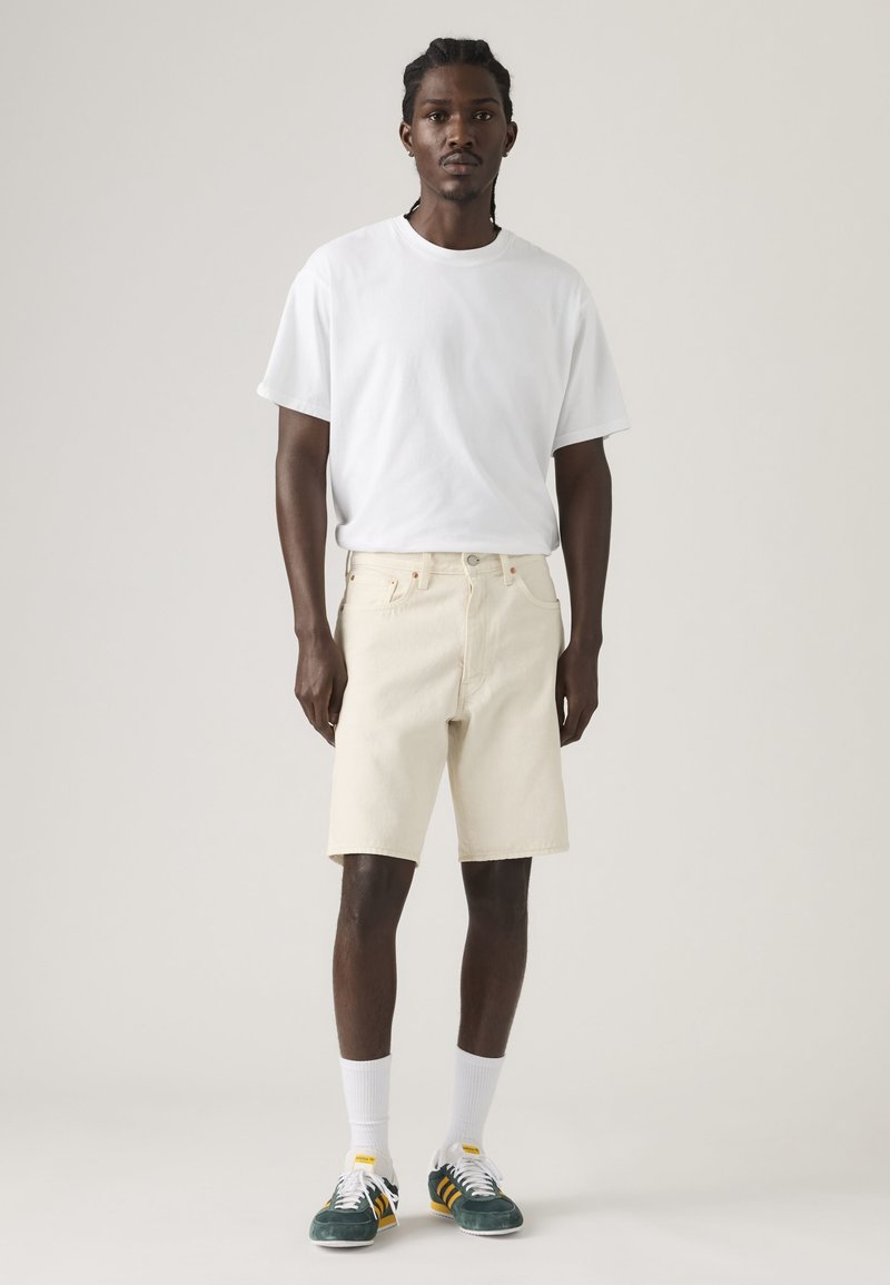 Uomo in piedi che indossa una t-shirt bianca semplice, pantaloncini beige fino al ginocchio, calzini bianchi e scarpe da ginnastica verde scuro davanti a uno sfondo neutro.