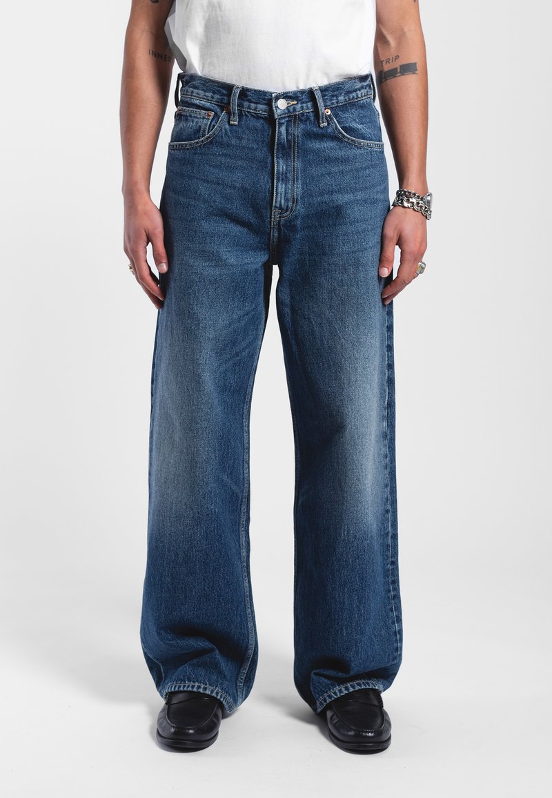 Wide-leg blue denim jeans featuring a classic five-pocket design, light fading, and visible stitching. Worn with a simple white shirt.