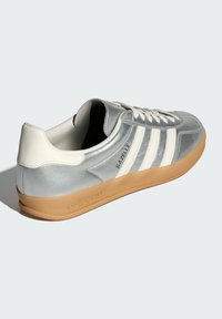 Silberne Sneakers mit weißen Akzenten, drei Streifen, einer strukturierten Oberfläche und einer Gummisohle. Das Logo an der Seite trägt die Aufschrift "Gazelle."