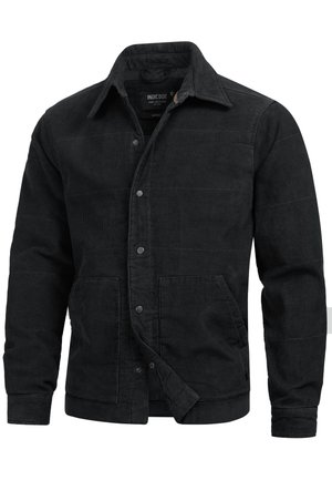 Smilende mand iført en sort kordfløjlsjakke med knapper og forlommer samt lyseblå jeans, stående mod en ensfarvet baggrund.