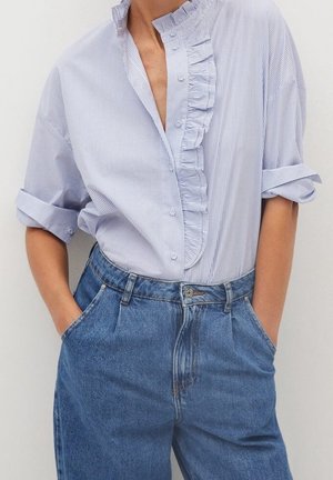 Personne portant un chemisier à rayures bleu clair et blanc avec un détail de volants et des manches retroussées, rentré dans un jean en denim bleu taille haute.