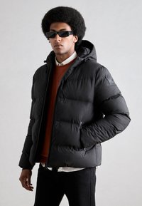 Czarna puchowa kurtka z kapturem, wyposażona w zamek błyskawiczny i kieszenie boczne. Teksturalny, pikowany wzór z subtelnym logotypem na ramieniu.