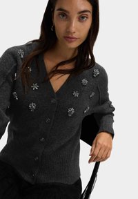 Ciemnoszary wełniany kardigan z dekoltem w serek, prążkowanym dołem, ozdobiony różnymi srebrnymi kwiatowymi detalami i zapięciem na guziki.