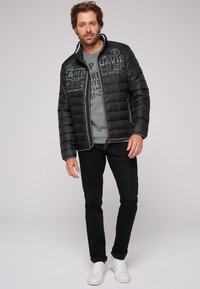 Giacca a vento nera con zip frontale, indossata sopra una felpa grigia con grafica, pantaloni neri e sneakers bianche.