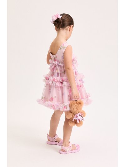 Νεαρή κοπέλα με φόρεμα ροζ με λουλούδια και σανδάλια, κρατώντας μια αρκουδάκι, με τα μαλλιά της πιασμένα σε κότσο διακοσμημένο με λουλούδια.