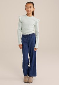 Svetlo modra dolga majica z nabrano rokavi, v kombinaciji z ohlapnimi temno modrimi kavbojkami in bež gležnjarji.