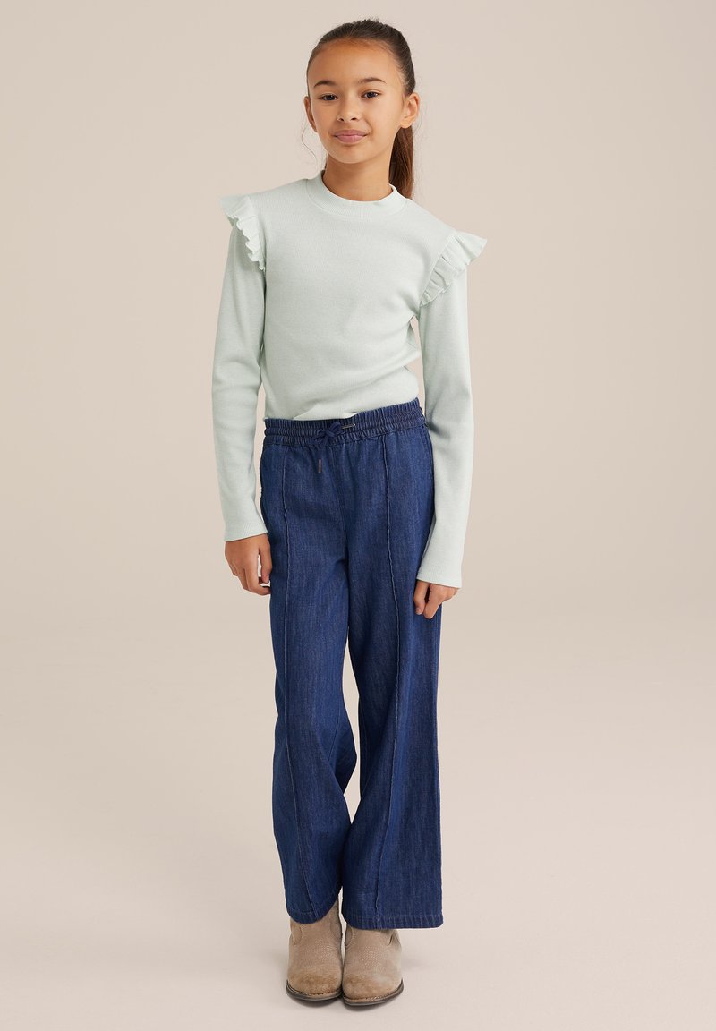 Svetlo modra dolga majica z nabrano rokavi, v kombinaciji z ohlapnimi temno modrimi kavbojkami in bež gležnjarji.