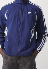 Navy blue zip-up jacket with white stripes along the sides and sleeves, featuring an Adidas logo on the chest and a collar.