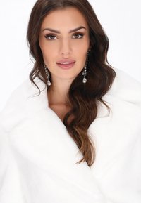 Donna con lunghi capelli castano mossi che indossa orecchini di perle pendenti e un morbido cappotto di pelliccia bianca su uno sfondo bianco.