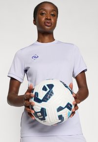 Lila, rövid ujjú sportpóló kerek nyakkivágással; egy fehér focilabdát tart, kék és narancssárga díszítésekkel és mintázatokkal.