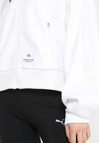 Veste blanche à fermeture éclair avec un ourlet côtelé, ornée d'une petite étiquette Calvin Klein. Associée à des leggings noirs arborant un logo Puma blanc.