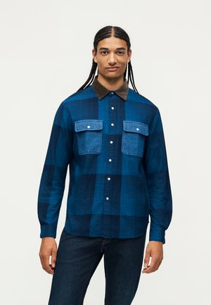 Young man with braided hair wearing a blue plaid button-up shirt with two chest pockets and dark blue jeans, standing against a plain background.