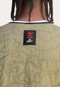 Kurtka w oliwkowym kolorze o pikowanym wzorze z kamuflażem, czarnymi wykończeniami i czerwoną metką Adidas na plecach w pobliżu dekoltu.