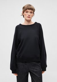 Melns, pārmēru liels džemperis ar plašu apaļu kakla izgriezumu un raglāna piedurknēm. Mīksta tekstūra un ribota aproce. Kombinēts ar tumšām, teksturētām biksēm.