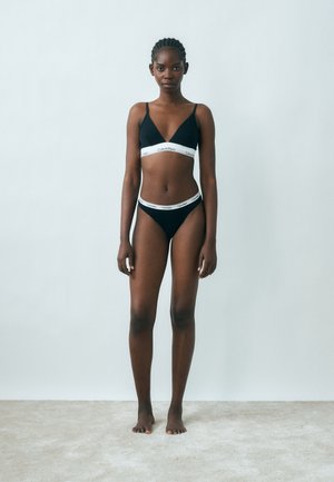 Žena stoji bosa, nosi crni Calvin Klein bralette i odgovarajući donji rublje, na jednostavnoj svijetlo sivoj pozadini.