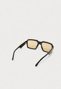 Óculos de sol quadrados pretos com lentes amarelas, armações de plástico espesso e hastes pretas elegantes com detalhes em dourado perto das dobradiças.