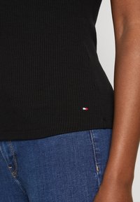 Top de tirantes negro acanalado con un diseño ajustado. Presenta un pequeño logo en rojo, blanco y azul en el dobladillo. Combinado con jeans de mezclilla.