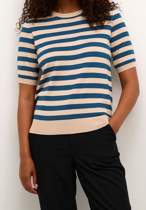 Vrouw draagt een beige en blauw horizontaal gestreept shirt met korte mouwen en zwarte broek, één hand in de zak, krullend haar zichtbaar vanaf de schouders naar beneden.